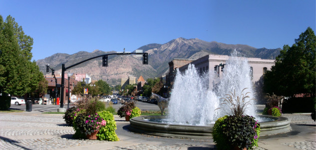 [Two photos stitched together showing the entire fountain (the painted horse is behind the water) and the metal pipe arch holding the street lights. The same buildings and mountains are in the background as the prior photo, but are not as prominent as this view focuses on the plants around the fountain as well as the fountain.]